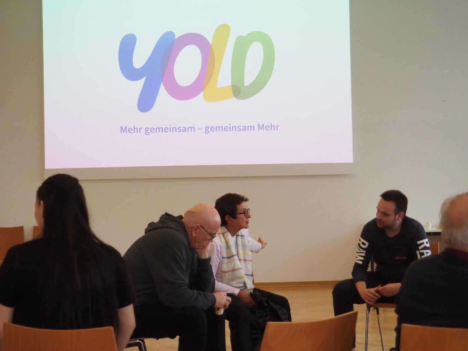 YOLD-Gruppenrunde: Jüngere und Ältere sitzen im Stuhlkreis, auf der Leinwand das YOLD-Logo
